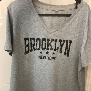 Grå t-shirt med Brooklyn-tryck - Snygg grå t-shirt med ett coolt Brooklyn New York-tryck i svart. T-shirten har en v-ringad hals och korta ärmar, perfekt för en avslappnad stil.