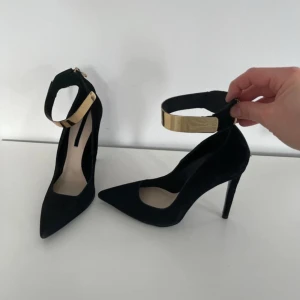 Svarta pumps med gulddetalj - Eleganta svarta pumps med spetsig tå och hög klack. Skorna har en stilren guldfärgad detalj runt ankeln som ger en lyxig känsla. Perfekta för en kväll ute eller en speciell tillställning.