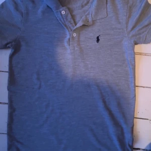 Grå pikétröja från Ralph Lauren - Säljer en klassisk grå pikétröja från Ralph Lauren med den ikoniska broderade logotypen på bröstet. Tröjan har korta ärmar och en stilren krage med knappar. Perfekt för en avslappnad men ändå stilfull look.