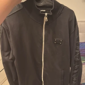 Svart jacka från Balmain - Snygg svart jacka från Balmain med dragkedja och en cool logotypdetalj på bröstet. Jackan har långa ärmar med Balmain-text längs sidan. Perfekt för en stilren look.