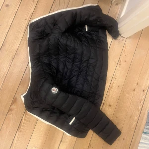 Svart dunjacka från Moncler - Säljer en snygg svart dunjacka från Moncler. Jackan har en quiltad design med dragkedja framtill och långa ärmar. Perfekt för kyligare dagar och ger en stilren look. Den är i storlek s men sitter bra på mig som även har m ibland. Jackan har väldigt fina vita detaljer. 