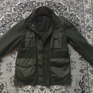 Grön jacka från J.Lindeberg - Snygg grön jacka från J.Lindeberg med flera praktiska fickor och en stilren design. Jackan har en dragkedja och knappar framtill samt en avtagbar huva. Perfekt för höst och vår. Det är S i storlek men den passar en M L
