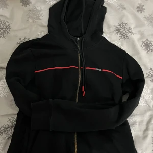 Svart hoodie från Hugo - Snygg svart hoodie från Hugo med dragkedja och röd detalj över bröstet. Perfekt för en casual look med sin bekväma passform och huva. Ett stilrent val för alla tillfällen.