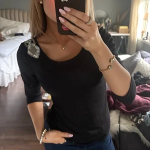 Svart topp med paljettdetaljer - Snygg svart topp med glittrande paljettdetaljer på axlarna och öppen rygg. Perfekt för en kväll ute eller en festlig tillställning. Toppen har trekvartsärmar och en figurnära passform.