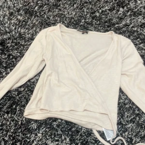Beige omlottopp - Säljer en stilren beige omlottopp med långa ärmar. Perfekt för en elegant och avslappnad look. Toppen har en knytning i sidan som ger en fin detalj. Passar bra till både jeans och kjol.