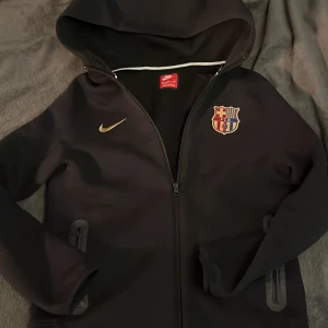 Svart Nike hoodie med FC Barcelona-logga - Säljer en svart hoodie från Nike med FC Barcelona-logga på bröstet. Perfekt för ett barcelona fan. Den är stor i storleken så jag skulle säga atta den passar 170-180cm långa. Den har inga tecken alls på användning och är fof mjuk och skön!