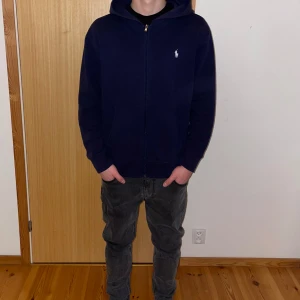 Mörkblå hoodie från Ralph Lauren - Säljer en stilren mörkblå hoodie från Ralph Lauren med dragkedja och broderad logga på bröstet. Tröjan har långa ärmar och en praktisk huva. Perfekt för en avslappnad look. Använd bara fåtal gånger! Dragskosnören tog jag bort. Nypris 2195kr