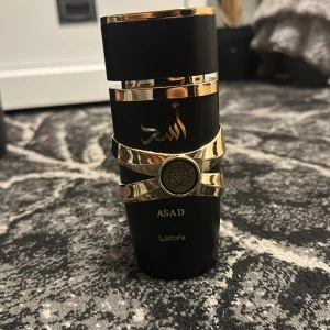 Asad parfym från Lattafa - Elegant parfymflaska i svart med gyllene detaljer från Lattafa. Flaskan har en lyxig design med guldfärgade band och en dekorativ medaljong. Perfekt för den som söker en sofistikerad doftupplevelse.