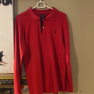 Röd pikétröja Ralph Lauren - Säljer en klassisk röd pikétröja från Ralph Lauren med långärmad design. Tröjan har en liten broderad logotyp på bröstet och en stilren krage med knappar. Perfekt för en avslappnad men stilren look, Passar som M