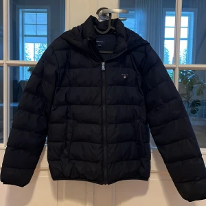 Svart pufferjacka från Gant - Säljer en snygg svart pufferjacka från Gant. Jackan har en dragkedja framtill och är långärmad med en liten logotyp på bröstet. Perfekt för kyligare dagar. Passar ungdomar i åldern 13-14 år.