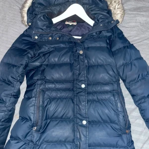 Mörkblå dunjacka från Tommy Hilfiger - Säljer en snygg mörkblå dunjacka från Tommy Hilfiger med avtagbar huva och fuskpäls. Jackan har dragkedja och knappar framtill samt praktiska fickor med dragkedja. Perfekt för kyliga dagar!