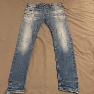 Blå jeans från Diesel - Säljer ett par snygga blå jeans från Diesel med en klassisk femficksdesign och slitna detaljer. De har en straight passform och är perfekta för en avslappnad stil. Midjan är normalhög och de har en knappgylf.