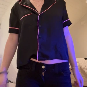 Sov tröja - Snygg svart blus med korta ärmar och rosa kantdetaljer. Blusen har knappar framtill och en klassisk krage. Perfekt för en avslappnad look.