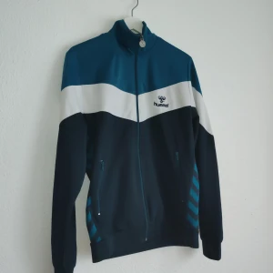 Blå och vit träningsjacka från Hummel - Säljer en snygg träningsjacka från Hummel i blått och vitt. Jackan har en dragkedja framtill och långa ärmar. Perfekt för sportiga aktiviteter eller som en stilren vardagsjacka.
