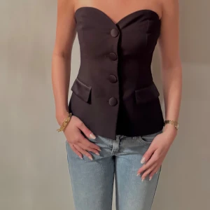 Svart tubtopp från Zara - Snygg svart tubtopp från Zara med stora knappar framtill och två dekorativa fickor. Perfekt för en stilren look. Toppen är axelbandslös och har en figurnära passform.