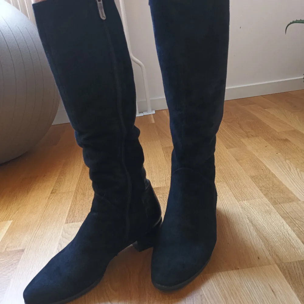 Snygga svarta stövlar i mocka med krokodilmönstrade detaljer och dragkedja på sidan.vinterstövlar i äkta läder med äkta päls inuti. Perfekta för en stilren look. De har en låg klack och är knähöga, vilket ger en elegant touch. . Kengät.