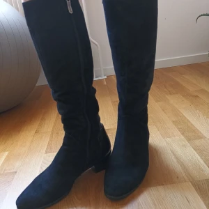 Svarta stövlar med krokodilmönster - Snygga svarta stövlar i mocka med krokodilmönstrade detaljer och dragkedja på sidan.vinterstövlar i äkta läder med äkta päls inuti. Perfekta för en stilren look. De har en låg klack och är knähöga, vilket ger en elegant touch. 