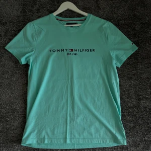 Turkos t-shirt från Tommy Hilfiger - Säljer en snygg turkos t-shirt från Tommy Hilfiger. Den har en klassisk design med märkesloggan tryckt på framsidan. Perfekt för en avslappnad stil och passar bra till både jeans och shorts. T-shirten är i storlek L och är gjord av mjuk bomull. Perfekt för sommardagar!