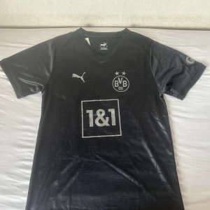 Svart Borussia Dortmund fotbollströja - Säljer en snygg svart Borussia Dortmund fotbollströja från Puma. Den har korta ärmar och är i bra skick. Tröjan har klubbens emblem och sponsorlogga på framsidan, samt spelarens namn och nummer 'Santos 22' på baksidan. Perfekt för alla BVB-fans!