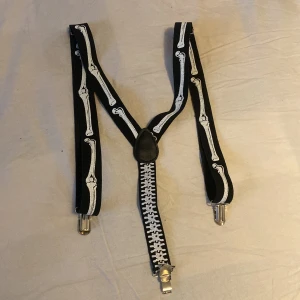 Svarta hängslen med skelettmönster - Coola svarta hängslen med ett unikt skelettmönster i vitt. Perfekta för att ge din outfit en edgy touch. De har metallklämmor för att enkelt fästa i byxorna. Passar perfekt för en fest eller Halloween!