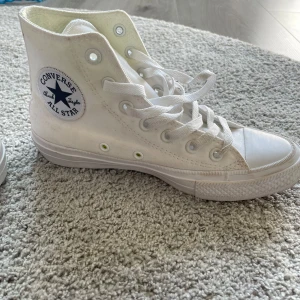 Vita Converse All Star sneakers - Säljer ett par klassiska vita Converse All Star sneakers i hög modell. Skorna har den ikoniska loggan på sidan och är i ny skick med vita snören och en gummisula. Perfekta för en avslappnad stil och passar till det mesta i garderoben. 