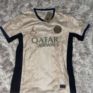 Beige och svart fotbollströja från Jordan - Säljer en snygg fotbollströja från Jordan i beige med svarta detaljer. Tröjan har korta ärmar och ett diskret mönster över hela tyget. Den har loggan för Qatar Airways på framsidan och en elegant Jordan-logga i guld. Perfekt för fotbollsfans som vill ha något unikt!