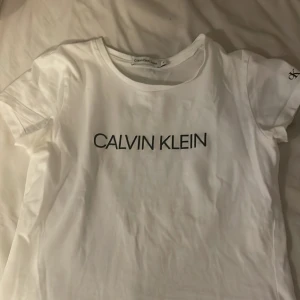 Vit t-shirt från Calvin Klein Jeans - Säljer en stilren vit t-shirt från Calvin Klein Jeans med svart logotyp på framsidan och texten 'JEANS' på baksidan. T-shirten har korta ärmar och är tillverkad i mjukt bomullsmaterial. Perfekt för en avslappnad look.