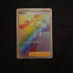 Pokémonkort - Worker - Säljer ett holografiskt Pokémonkort med karaktären Worker. Kortet är en Supporter-kort och har en färgglad, skimrande yta. Perfekt för samlare eller spelare som vill förstärka sin kortlek. Kortet har en klassisk Pokémon-baksida med en Pokéball.