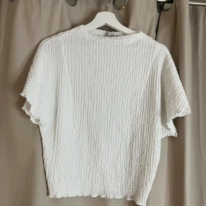 Vit topp från Zara - Säljer en vit topp från Zara med en texturerad yta. Toppen har korta ärmar och en avslappnad passform. Perfekt för en stilren look.