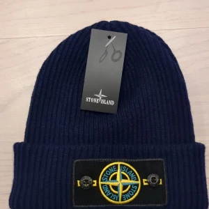 Mörkblå ribbad mössa från Stone Island - Snygg Stone island mössa med ribbad design och uppvikt kant. Mössan har en broderad logotyp framtill och är perfekt till kallare dagar. Skriv vid minsta fråga.