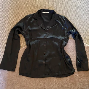 Svart skjorta - Elegant svart blus i siden från Lager 157 med klassisk krage och knappar framtill. Perfekt för en stilren look. Blusen har långa ärmar och en mjuk, glansig finish som ger en lyxig känsla.😻Är gjord för sovplagg men du kan ha den till vad som helst, med jeans utomhus t.ex.❤️Den är så mjuk, luftig och skön med tanke på silkes materialet💕Du får med en extra knapp💋