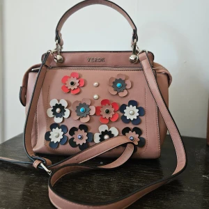 Beige axelväska med blommor från Verde - Snygg beige axelväska från Verde dekorerad med färgglada blommor i olika nyanser. Väskan har både handtag och en justerbar axelrem. Tillverkad i syntetmaterial med polyesterfoder. Perfekt för att ge en färgklick till din outfit!