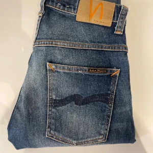 Blå jeans från Nudie Jeans - Snygga blå jeans från Nudie Jeans med klassisk femficksdesign och orange sömmar. Jeansen har en slim passform och är tillverkade i slitstarkt denim. Perfekta för en avslappnad stil.