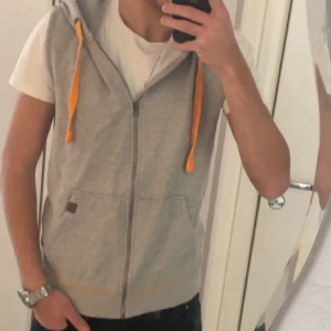 Grå ärmlös huvtröja med dragkedja - Säljer en grå ärmlös huvtröja med dragkedja och orangea snören. Perfekt för en avslappnad stil. Den har en praktisk ficka framtill och en bekväm huva. Den är använd ett par gånger maximalt 6 gånger, pris går såklart att diskuteras, och vid andra frågor så är det bara att skriva! ABS! Den fungerar lika bra om man har S då jag har själv S i vissa kläder och den sitter väldigt bra.