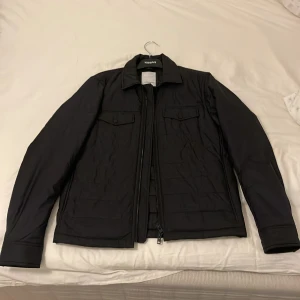 Svart quiltad jacka från J.Lindeberg - Säljer en stilren svart quiltad jacka från J.Lindeberg. Jackan har en dragkedja framtill och två bröstfickor med knappar. Perfekt för kyligare dagar med sin snygga och funktionella design.