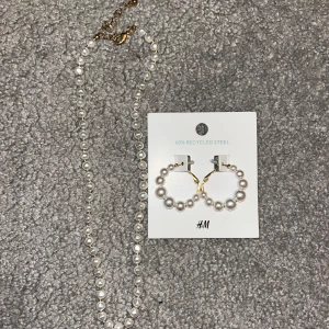 Pärlhalsband och örhängen från H&M - Elegant pärlhalsband och matchande örhängen från H&M. Halsbandet har små vita pärlor och en justerbar kedja i guldton. Örhängena är cirkulära med vita pärlor och guldfärgade detaljer. Både halsbandet och öronhänherna följer med i beställningen! Aldrig använd