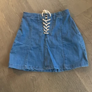 Blå jeanskjol med snörning - Snygg blå jeanskjol från Perfect Jeans med cool snörning framtill. Kjolen har en kort längd och en klassisk jeanslook som passar perfekt till många olika stilar. Perfekt för dig som vill ha en trendig och bekväm kjol i garderoben.