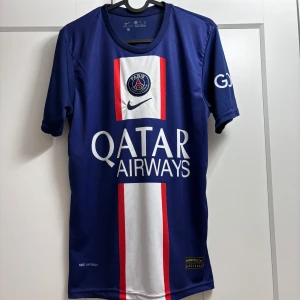 Blå PSG fotbollströja - Snygg blå PSG fotbollströja från Nike med Neymar Jr och nummer 10 på ryggen. Tröjan har korta ärmar och ett vitt och rött vertikalt mönster på framsidan. Perfekt för fans av Paris Saint-Germain och Neymar!