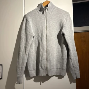 Grå stickad kofta från Selected Homme - Säljer en stilren grå stickad kofta från Selected Homme. Koftan har en dragkedja framtill och en hög krage som ger extra värme. Perfekt för kyligare dagar. Passar bra till både jeans och chinos. Perfekt nu till våren. Skick 7/10, tröjan är sparsamt använd och inga större tecken på användning 