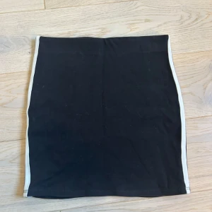 Svart kjol med vita och gula ränder - Snygg svart kjol med vita och gula ränder längs sidorna. Perfekt för en sportig look. Kjolen är kort och har en elastisk midja för bekväm passform. Köpt på barnavdelningen men passar som en xs/s och är samt väldigt stretchig. Använd men är som ny💛🖤