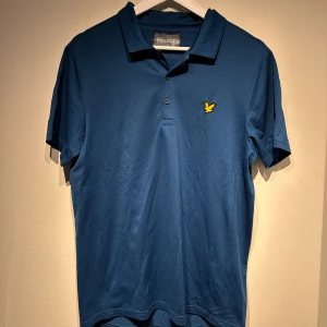 Blå pikétröja från Lyle & Scott - Snygg blå pikétröja från Lyle & Scott med korta ärmar och klassisk krage. Tröjan har en gul logotyp på bröstet och knappar framtill. Perfekt för en stilren look.
