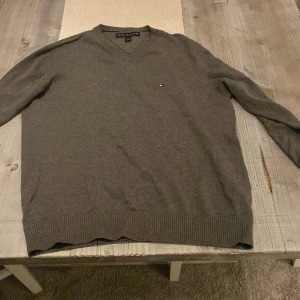 Grå tröja från Tommy Hilfiger - Säljer en stilren grå tröja från Tommy Hilfiger med v-ringning och diskret logga på bröstet. Tröjan har långa ärmar och ribbade muddar vid ärmslut och nederkant. Perfekt för en avslappnad och snygg look.