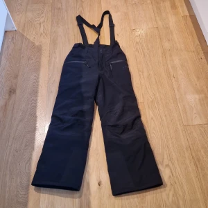 Svarta täck/skidbyxor från McKinley - Svarta täck/skid byxor från McKinley med justerbara hängslen och dragkedjor på sidorna. Perfekta för vinterns kalla dagar. Byxorna har en stilren design med logotyp på benet. Toppskick. Storlek:146. Som nyskick 