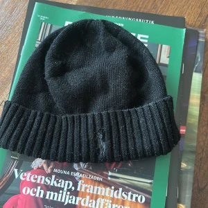 Svart stickad mössa från Ralph Lauren - Säljer en klassisk svart stickad mössa från Ralph Lauren med ribbad kant och det ikoniska broderade logotypen i vitt. Perfekt för kyliga dagar. Använd men väldigt bra slock