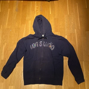 Mörkblå zip hoodie från Von Studio - Säljer en mörkblå zip hoodie från Von Studio med glittrande text på framsidan och ett tryck på baksidan på baksidan. Storlek S, 700kr nypris. Ett litet hål längst ner i armen som man inte tänker på.