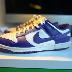 Nike Dunk i lila och vitt - Snygga Nike Dunk sneakers i en fräsch kombination av lila och vitt. Skorna har klassisk snörning och en stilren design med det ikoniska Nike-märket på sidan. Perfekta för en sportig look. Har använts varsamt och med creese skydd.