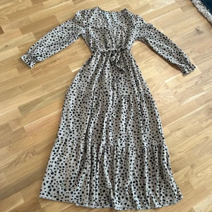 Beige prickig långklänning - Snygg beige långklänning med svarta prickar och knytband i midjan. Klänningen har långa ärmar och en v-ringad design som ger en elegant look. Perfekt för en stilfull dag eller kväll.