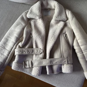 Beige jacka från Stradivarius - Säljer en snygg beige jacka från Stradivarius med mjukt foder och stiliga dragkedjedetaljer. Jackan har en bälte i midjan och är perfekt för kyligare dagar. Den har en klassisk design med en modern twist.