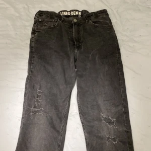 Svarta skinny jeans från &Denim - Snygga svarta skinny jeans från &Denim med slitna detaljer på benen. De har en knapp och dragkedja framtill samt fem fickor. Byxorna har även resår i midjan så dom är ställbara om dom skulle va lite för stora. Även ett fodrat lager på insidan som gör dom lite varmare än vanliga jeans! 