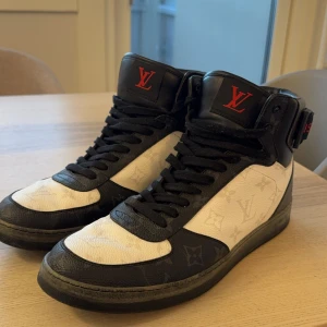 Svartvita sneakers från Louis Vuitton - Snygga svartvita sneakers från Louis Vuitton med röd logotypdetalj. Skorna har snörning och ett klassiskt monogrammönster. Perfekta för den stilmedvetna som vill ha något unikt. 
